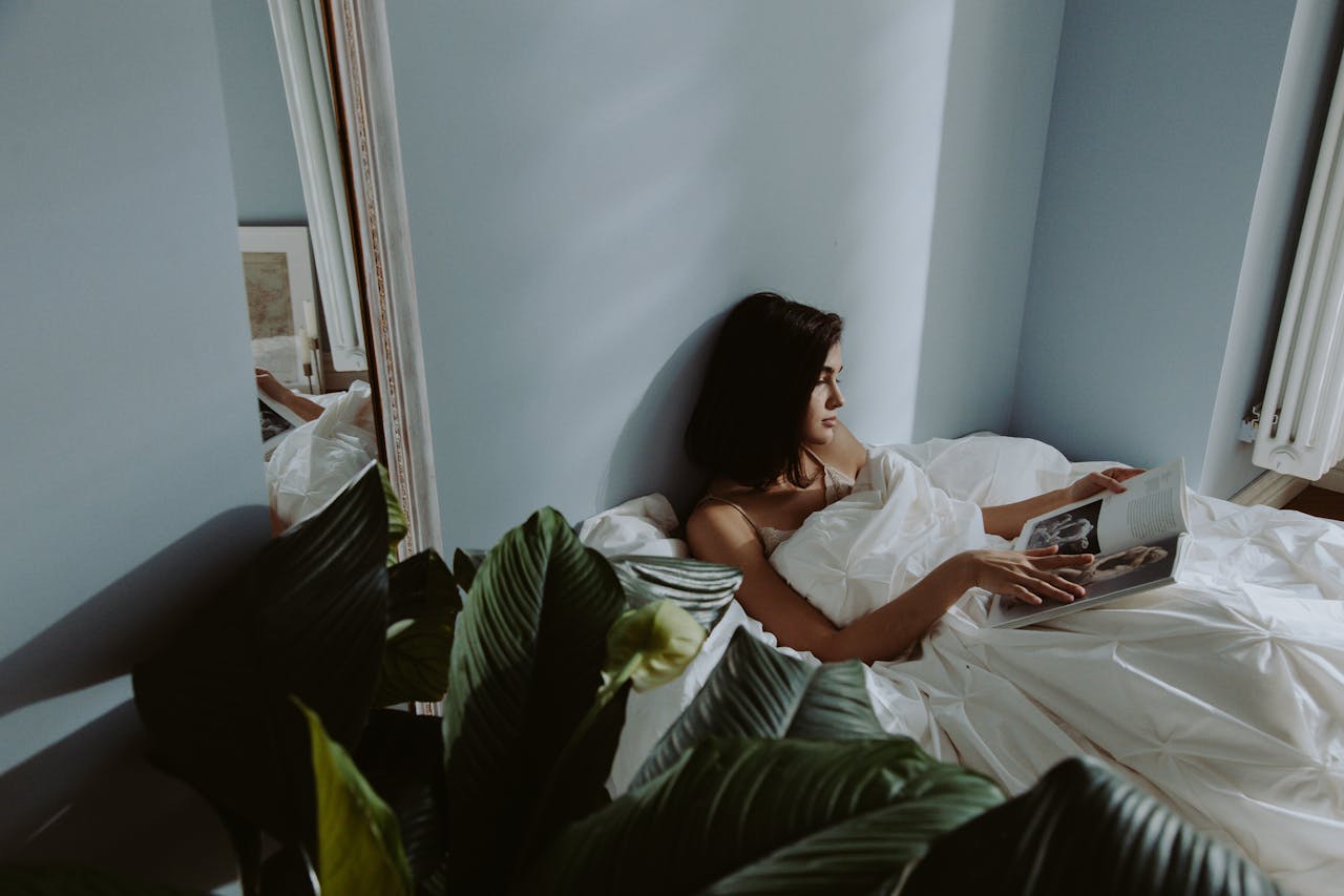 about-img-01 A woman reads a book in bed under morning light, surrounded by plants and reflections.