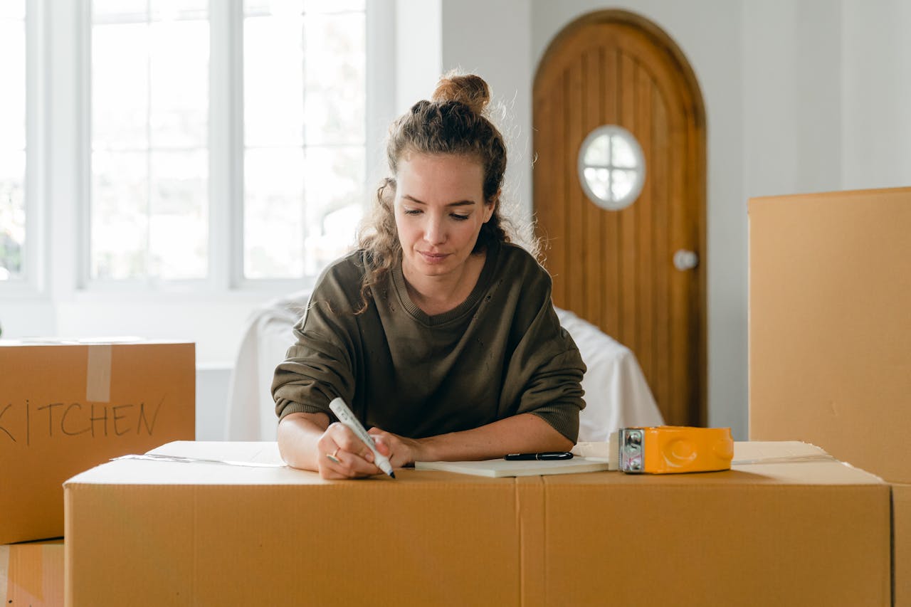 gallery-04 A woman labels cardboard boxes while preparing to move, with sunlight streaming through windows.